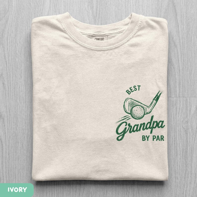 May include: An ivory short-sleeved t-shirt featuring a green graphic on the chest. The design includes a golf club and ball with the text "BEST" above and "Grandpa BY PAR" below the illustration, printed in a dark green ink on the fabric.