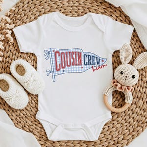 May include: White baby bodysuit featuring a blue and red "Cousin Crew" flag design. The flag has a gingham pattern and the name "Liam" in red script. A pair of white baby shoes and a crochet bunny rattle are next to the bodysuit.