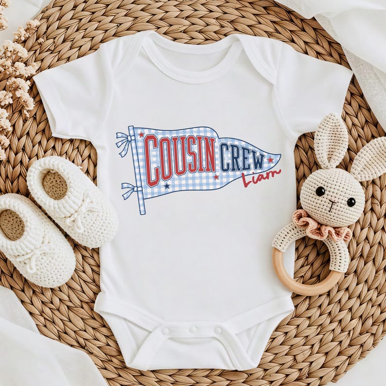 May include: White baby bodysuit featuring a blue and red "Cousin Crew" flag design. The flag has a gingham pattern and the name "Liam" in red script. A pair of white baby shoes and a crochet bunny rattle are next to the bodysuit.