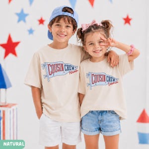 May include: Two children wearing matching beige t-shirts with a "Cousin Crew" flag design. The flag has a blue and white gingham pattern and the names "Noah" and "Sophia" are printed on the shirts. The background has red, white, and blue star decorations.
