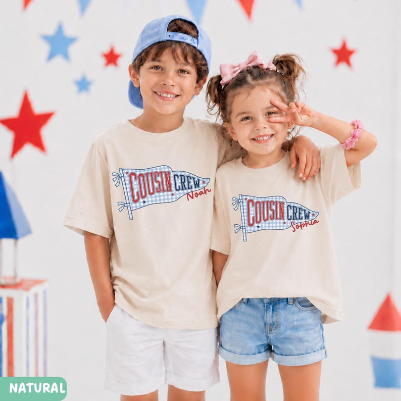 May include: Two children wearing matching beige t-shirts with a "Cousin Crew" flag design. The flag has a blue and white gingham pattern and the names "Noah" and "Sophia" are printed on the shirts. The background has red, white, and blue star decorations.