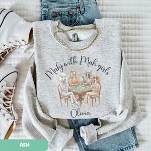 Op de afbeelding: Asgrijze sweatshirt met de tekst "Mahj with Mah girls" en een afbeelding van honden die mahjong spelen. De naam "Olivia" staat onder de afbeelding. Een gouden ketting en een blauwe jeans zijn ook zichtbaar.