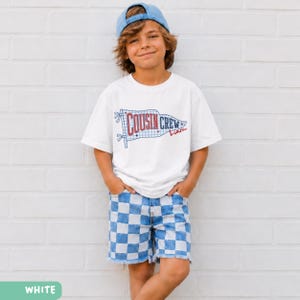 May include: A white t-shirt featuring a "Cousin Crew" flag design and a name in red script. The outfit includes blue and white checkered shorts and a blue baseball cap. The clothing is worn by a young person.