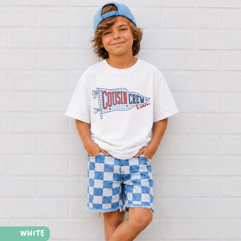 May include: A white t-shirt featuring a "Cousin Crew" flag design and a name in red script. The outfit includes blue and white checkered shorts and a blue baseball cap. The clothing is worn by a young person.
