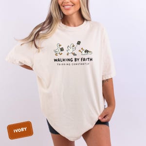 May include: Ivory-colored t-shirt featuring a whimsical illustration of ducks with Bibles, with the text "WALKING BY FAITH TRIPPING CONSTANTLY". The ducks are depicted in various poses, some upright and others stumbling.