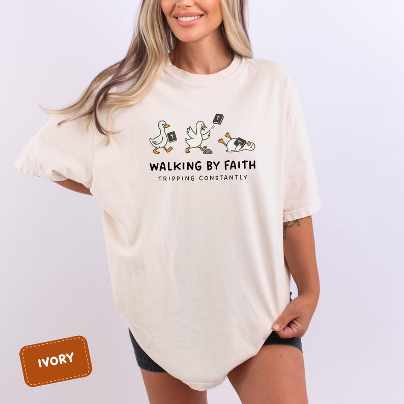 May include: Ivory-colored t-shirt featuring a whimsical illustration of ducks with Bibles, with the text "WALKING BY FAITH TRIPPING CONSTANTLY". The ducks are depicted in various poses, some upright and others stumbling.