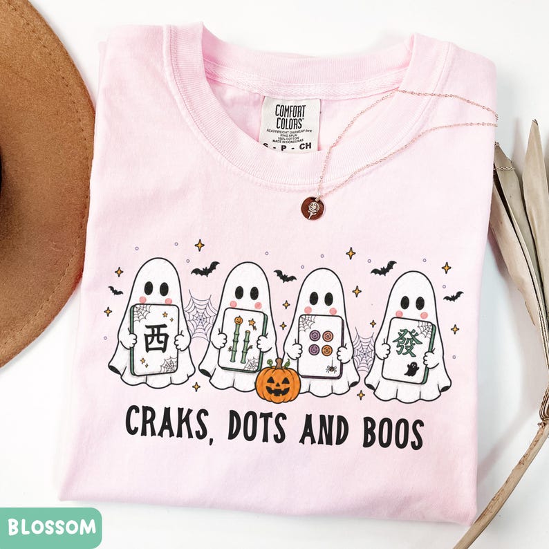 May include: A light pink t-shirt featuring a graphic of four white ghosts holding mahjong tiles, a small orange pumpkin, spiderwebs, bats, and stars. The text "CRAKS, DOTS AND BOOS" is printed in black below the ghosts. The shirt has a crew neckline.