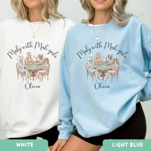 Op de afbeelding: Witte en lichtblauwe sweatshirts met een afbeelding van vrouwen die een kaartspel spelen. De tekst "Mahy with Mah girls" en de naam "Olivia" staan op de voorkant. Het lichtblauwe sweatshirt is onderaan voorzien van het label "LIGHT BLUE".
