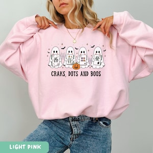 May include: A light pink crewneck sweatshirt featuring a graphic of four white ghosts holding mahjong tiles, a small orange jack-o'-lantern, spiderwebs, and bats. The text below the ghosts reads, "CRAKS, DOTS AND BOOS" in black lettering.