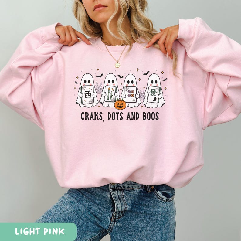 May include: A light pink crewneck sweatshirt featuring a graphic of four white ghosts holding mahjong tiles, a small orange jack-o'-lantern, spiderwebs, and bats. The text below the ghosts reads, "CRAKS, DOTS AND BOOS" in black lettering.