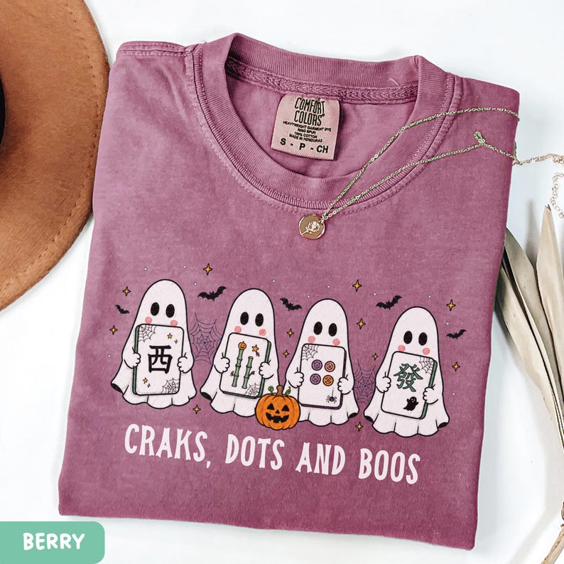 May include: A berry-colored t-shirt featuring a graphic of four white ghosts holding mahjong tiles, a small orange pumpkin, spiderwebs, and bats. The text reads "CRAKS, DOTS AND BOOS" in white capital letters. The shirt has a Comfort Colors brand label.