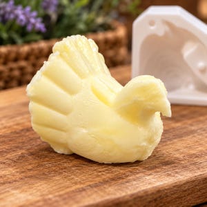 May include: A pale yellow butter sculpture shaped like a turkey with a fanned tail and defined wing details, positioned next to a white silicone mold on a wooden surface. The turkey figure displays a smooth, molded texture.