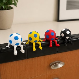 May include: Four small soccer ball figurines with articulated arms and legs, each sitting on a ledge. The balls are colored white and blue, yellow and blue, red and blue, and black and white. Each figure has matching colored limbs and feet.