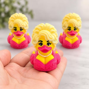 Könnte beinhalten: Drei gelbe Quietscheentchen mit blondem Haar, die leuchtend pinke, strukturierte Kleider und weiße Perlenketten tragen. Jede Ente hat dunkle Wimpern, betonte Augenbrauen und einen leuchtend pinken Schnabel als kleine Sammelfigur gestaltet.