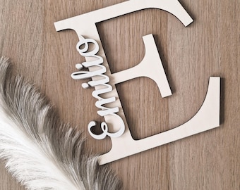 Op de afbeelding: Een houten letter "E" met de naam "Emilie" in cursief schrift, tegen een achtergrond van houtnerf. Een decoratieve witte veerkwast bevindt zich in de linker benedenhoek. Dit item is geschikt voor woondecoratie.