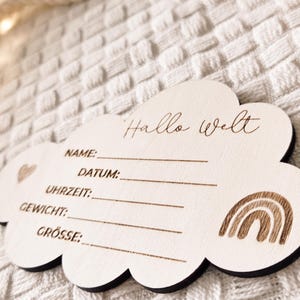 Op de afbeelding: Een houten wolk-vormig bord met de woorden "Hallo Welt" en lijnen voor Naam, Datum, Tijd, Gewicht en Grootte. Een hart en een regenboog zijn op het bord geëtst. Het bord ligt op een wit, getextureerd oppervlak.
