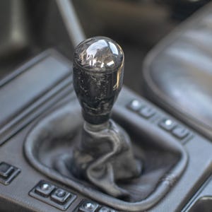 May include: A close-up shot of a car's gear stick. The gear knob is dark grey with a textured pattern, reflecting light. The shift boot is black leather, and the console is dark grey with buttons.