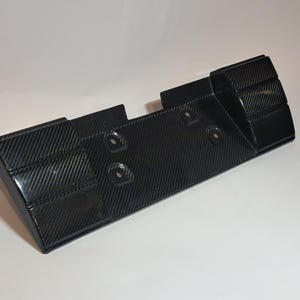Puede incluir: Un componente automotriz de fibra de carbono negra con un patrón de tejido texturizado. La pieza tiene forma rectangular con bordes redondeados y varios orificios de montaje. El diseño incluye secciones elevadas en los laterales.