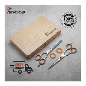 May include: A set of two silver-toned metal hair cutting shears with gold-colored finger rings and inserts, accompanied by a rectangular light-colored wooden storage box featuring the logo THE LINE OF ART QUALITY DOES MATTER.
