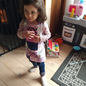 May include: A young girl wearing a pink and floral patchwork apron with the word "Grandma" embroidered on it. She is standing on a wooden floor with a toy kitchen in the background.