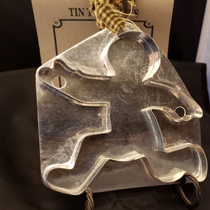 May include: A silver metal gingerbread man cookie cutter. The cutter is shaped like a gingerbread man with arms outstretched and legs spread apart. The cutter has a hole in the top for hanging.