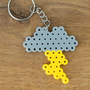 May include: A silver-colored cloud-shaped keychain with a yellow lightning bolt charm. The cloud has small, evenly spaced holes. The keychain is on a wooden surface.
