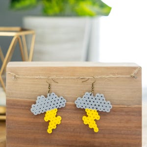 May include: A pair of gray and yellow cloud earrings with a lightning bolt design. The earrings are made of plastic beads and are hanging from a wooden surface.