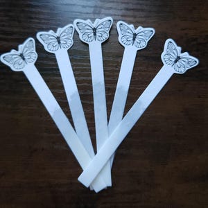 Può includere: Cinque segnalibri di carta bianca con un disegno a farfalla. Ogni segnalibro presenta un'illustrazione di farfalla in bianco e nero nella parte superiore, con un lungo stelo rettangolare. I segnalibri sono disposti a ventaglio.