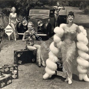 May include: Vintage black and white photograph featuring several women in glamorous attire posing near a classic car. One woman wears a sparkling gown and a large white fur wrap. Suitcases are in the foreground, and a "Curves Ahead" sign is visible.
