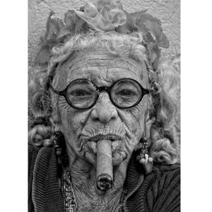 May include: A black and white portrait of an older woman with gray hair, wearing glasses and a flower crown, smoking a cigar.