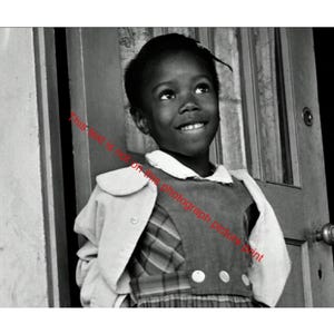 May include: Black and white photograph of a young girl smiling and looking upwards. She is wearing a collared dress with a vest and a light-colored jacket draped over her shoulders. The background includes a door and a wall.