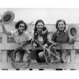 May include: Black and white photograph of three women smiling and holding cowboy hats and ropes. They are leaning on a wooden fence, dressed in western attire, and appear to be enjoying themselves. The image captures a vintage, rustic aesthetic.