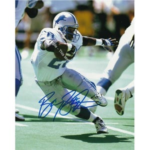 May include: A football player wearing a blue and white uniform with the number 62 on the front. He is running with the ball and has a determined expression on his face. The player is wearing a blue helmet with a lion logo. The image is signed by the player.