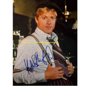 Op de afbeelding: Een portret van een man in een lichtblauw overhemd met bretels en een stropdas met patroon. Hij draagt ook een donkere hoed en heeft een handtekening met blauwe inkt op het overhemd.