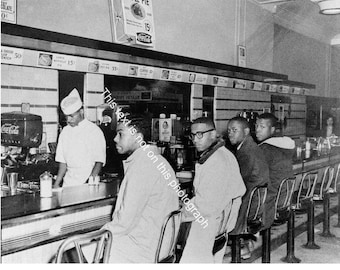 Puede incluir: Fotografía en blanco y negro de una escena de restaurante. Un camarero con uniforme blanco está detrás de un mostrador, sirviendo a los clientes. Varias personas están sentadas en el mostrador en taburetes de metal. Los elementos del menú y los precios son visibles encima del mostrador.
