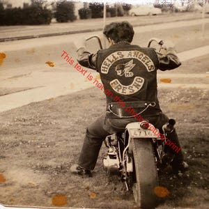 May include: Vintage black and white photograph of a person on a motorcycle. The person is wearing a leather jacket with a "HELLS ANGELS FRESNO" patch on the back. The motorcycle has a license plate that reads "47 16".