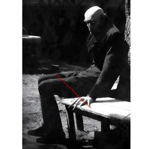 May include: Black and white photograph of a man in a dark coat and hat, seated on a wooden bench. The man has a bald head and a stern expression. The image is a vintage portrait, likely from a film or stage production.
