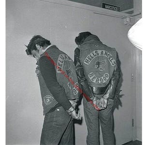 May include: Two individuals wearing denim vests and jackets with "HELLS ANGELS" patches on the back, standing in a room. The jackets have a circular emblem with a winged skull. The individuals are handcuffed. A sign above reads "NARCOTICS".