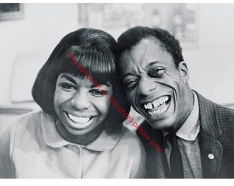 May include: Black and white photograph of two people smiling widely. The woman on the left has short, dark hair and is wearing a collared shirt. The man on the right is wearing a blazer and tie. Both are laughing.