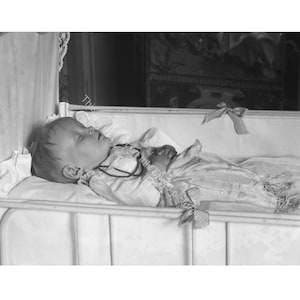 May include: A sleeping baby in a vintage crib. The baby is wearing a light-colored outfit and a necklace. The crib has a white metal frame and is lined with a white sheet and pillow. A decorative bow is tied to the crib.