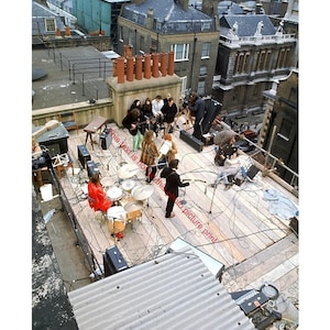 May include: A rooftop scene featuring a band performing, with a drum set, amplifiers, and microphones. The band members are dressed in various outfits. The setting includes buildings and chimneys in the background, with cables and equipment on the wooden platform.