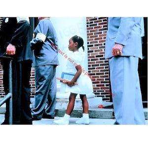May include: A young girl in a white dress and shoes stands between several adults in suits. She holds books and a binder. The background includes a brick wall and a white column. The image is a historical photograph.