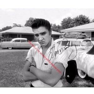 May include: Black and white photograph of Elvis Presley with his arms crossed, standing in front of a car and a house. The image includes his signature. The photo is taken outdoors on a sunny day.