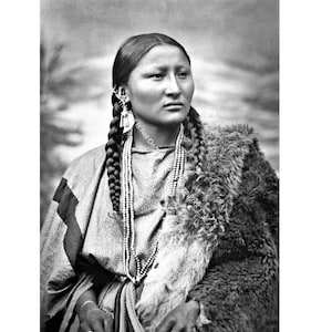 May include: Black and white portrait of a Native American woman wearing a fur coat and beaded necklaces. She has long braided hair and a serious expression.