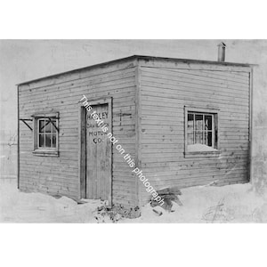 May include: A black and white photograph of a small wooden building covered in snow. The building has two windows and a door with the text "Harley Davidson Motor Co." The building has a chimney and is surrounded by snow.