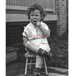 May include: A young child with curly hair sits on a wooden stool, wearing a white button-down shirt and shorts. They are holding a cigar in their hand and looking at the camera.