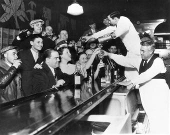 May include: Black and white vintage photograph of a bar scene. A bartender pours drinks for a crowd of people. The scene is filled with people, glasses, and bottles, capturing a moment of celebration.