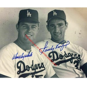 May include: Black and white photograph of two men wearing baseball caps and Dodgers jerseys. The jerseys are white with blue lettering and the Dodgers logo. The caps are black with a white "LA" logo. Both men are smiling.