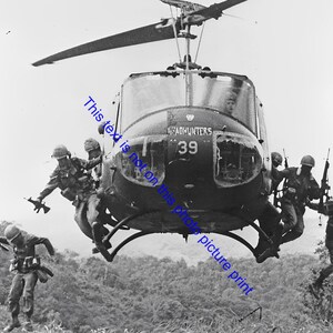 May include: Black and white photograph of soldiers exiting a helicopter. The helicopter, marked with "HEADHUNTERS 39", hovers above a forested landscape. Soldiers are in mid-air, some holding rifles.