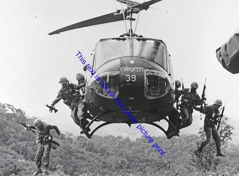 May include: Black and white photograph of soldiers exiting a helicopter. The helicopter, marked with "HEADHUNTERS 39", hovers above a forested landscape. Soldiers are in mid-air, some holding rifles.